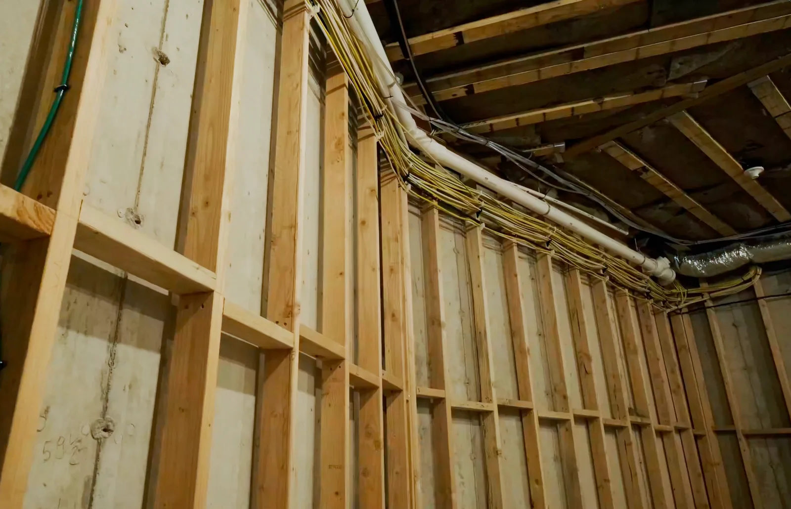 Interior of a house under construction at the framing stage — exposed wood wall studs with PVC piping and bundled electrical wiring running along the ceiling